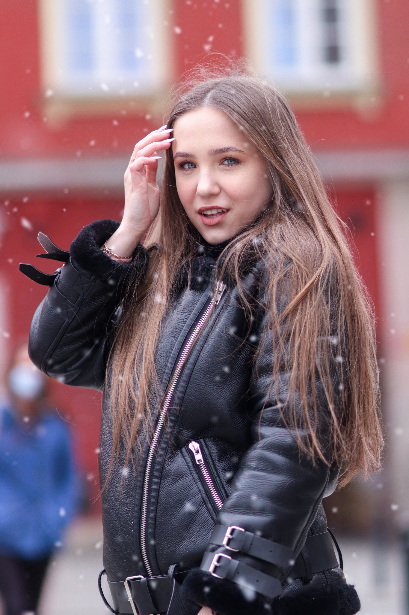 Portret młodej kobiety w czarnej kurtce z futerkiem, delikatny uśmiech, padający śnieg, czerwona ściana budynku w tle.