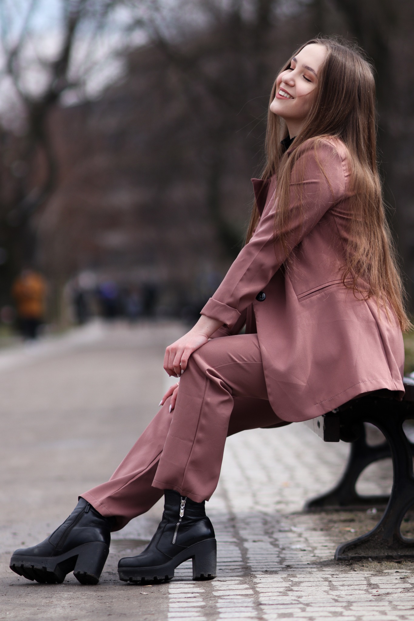 Uśmiechnięta młoda kobieta w różowym garniturze i czarnych botkach siedzi na ławce w parku, z głową odchyloną do tyłu i zamkniętymi oczami.