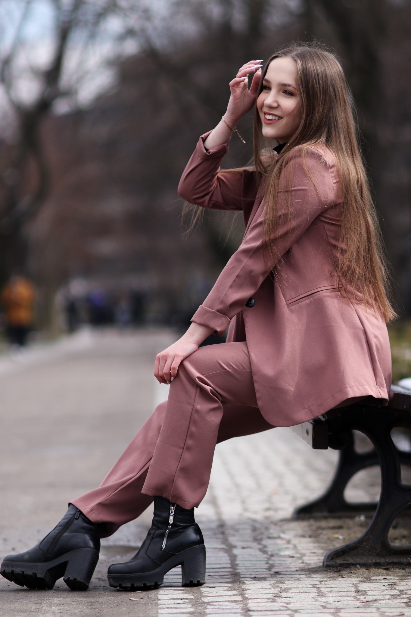 Uśmiechnięta młoda kobieta w różowym garniturze siedzi na ławce w parku, poprawiając włosy, z widocznymi czarnymi botkami na platformie.