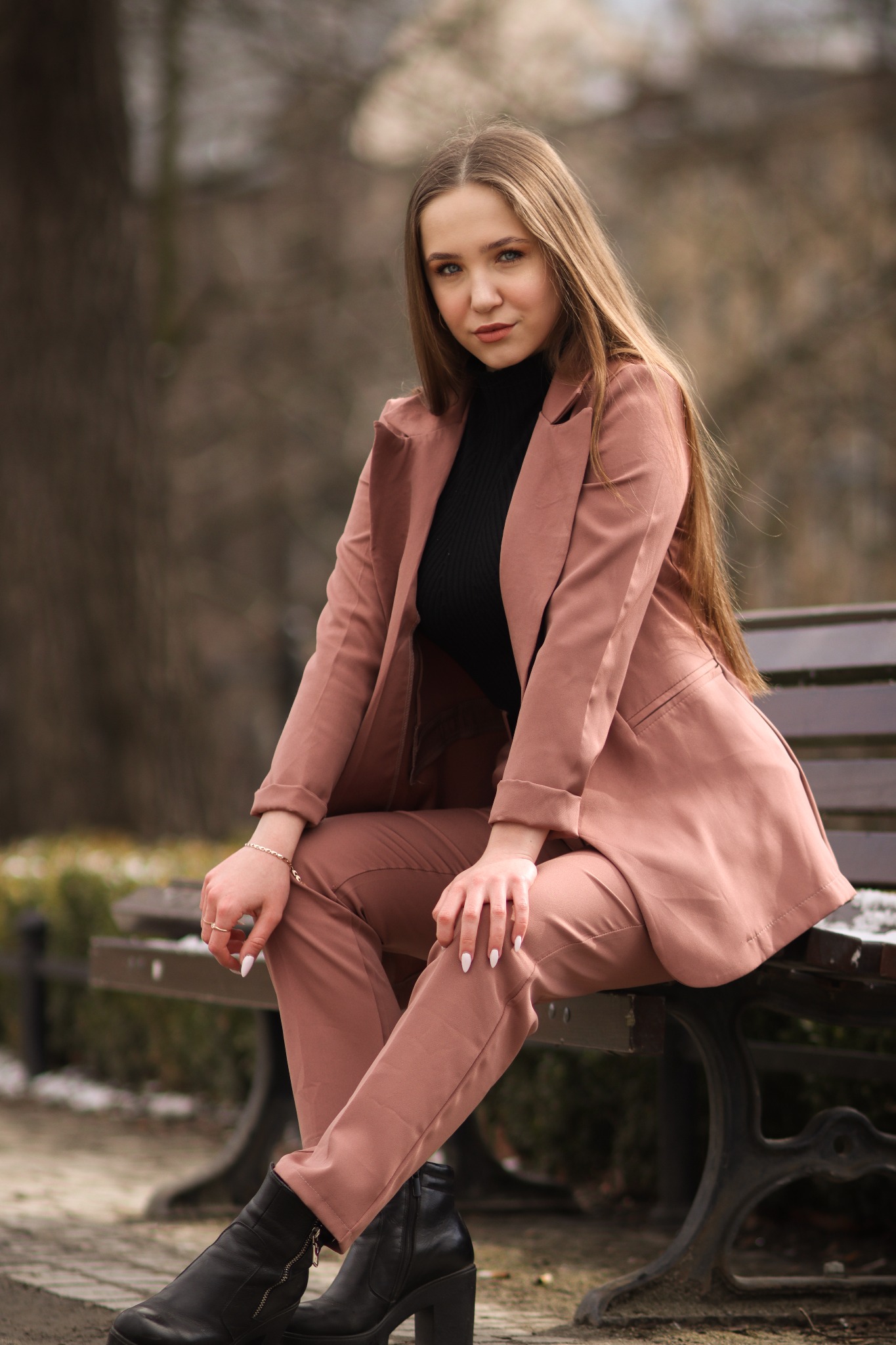 Młoda kobieta w różowym garniturze i czarnym golfie siedzi na ławce w parku, pozując do portretu z delikatnym uśmiechem.