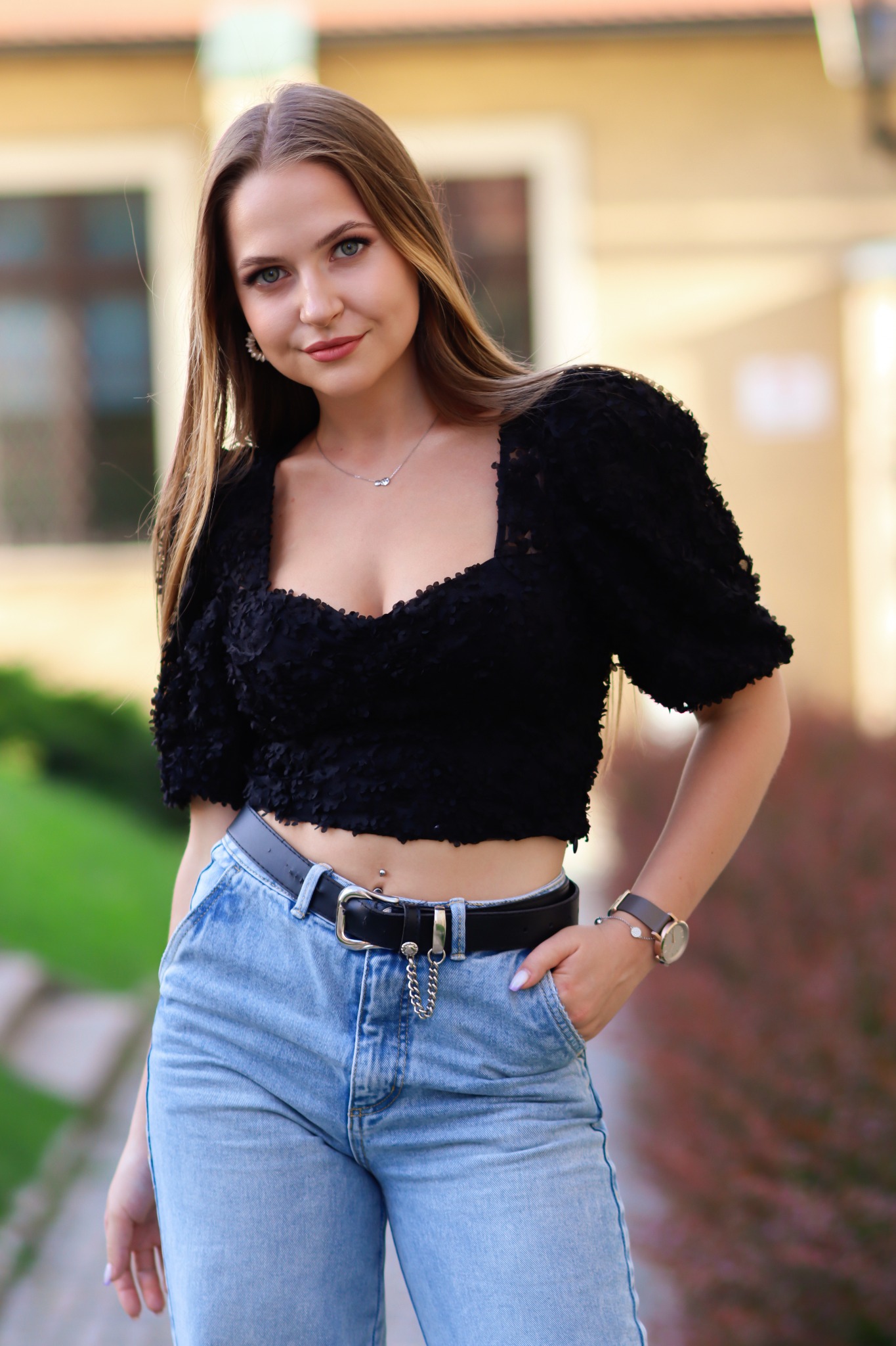 Portret młodej kobiety w czarnej bluzce z bufiastymi rękawami i jasnych jeansach z paskiem, stojącej na tle budynku z zielenią.