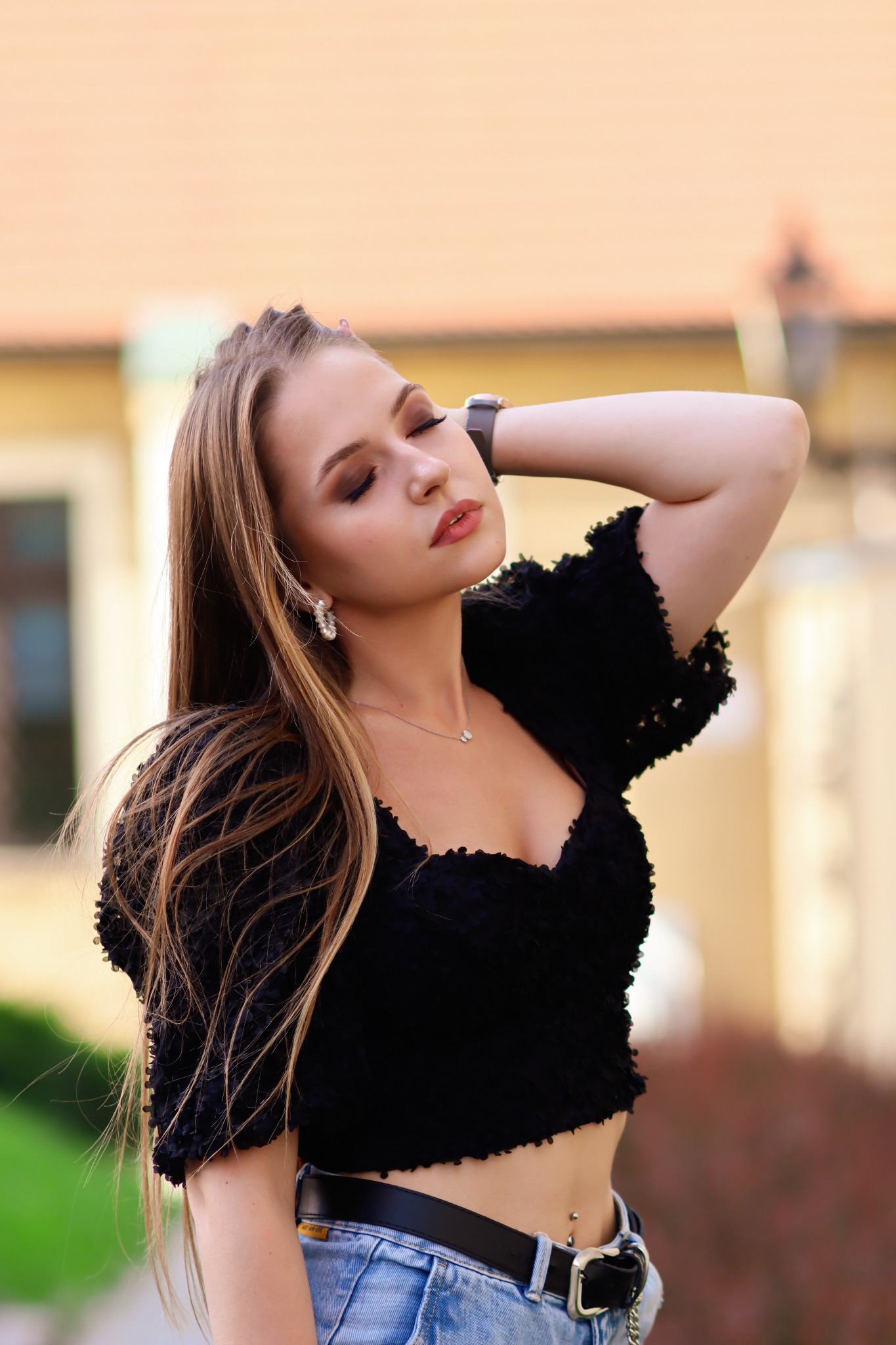 Portret młodej kobiety z długimi włosami, w czarnej bluzce i jeansach, pozującej na tle budynku z cegły.