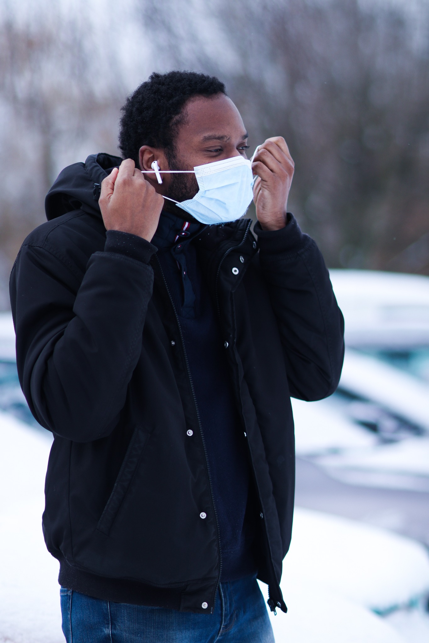 Mężczyzna w czarnej kurtce i jeansach, poprawia maseczkę ochronną na twarzy, widoczne słuchawki bezprzewodowe w uszach, zima w tle.