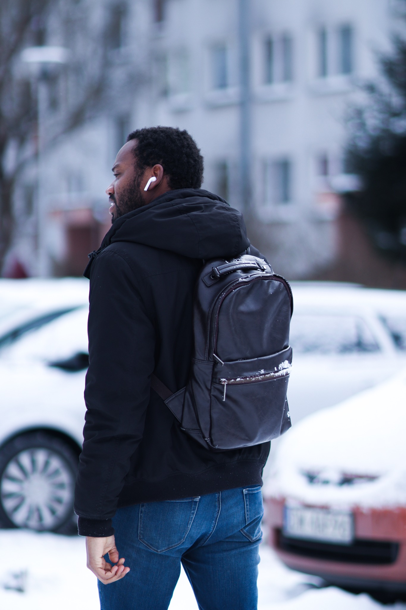 Mężczyzna z plecakiem idzie zimą, widoczny z profilu, w słuchawkach bezprzewodowych, na tle zaśnieżonych samochodów i budynku.