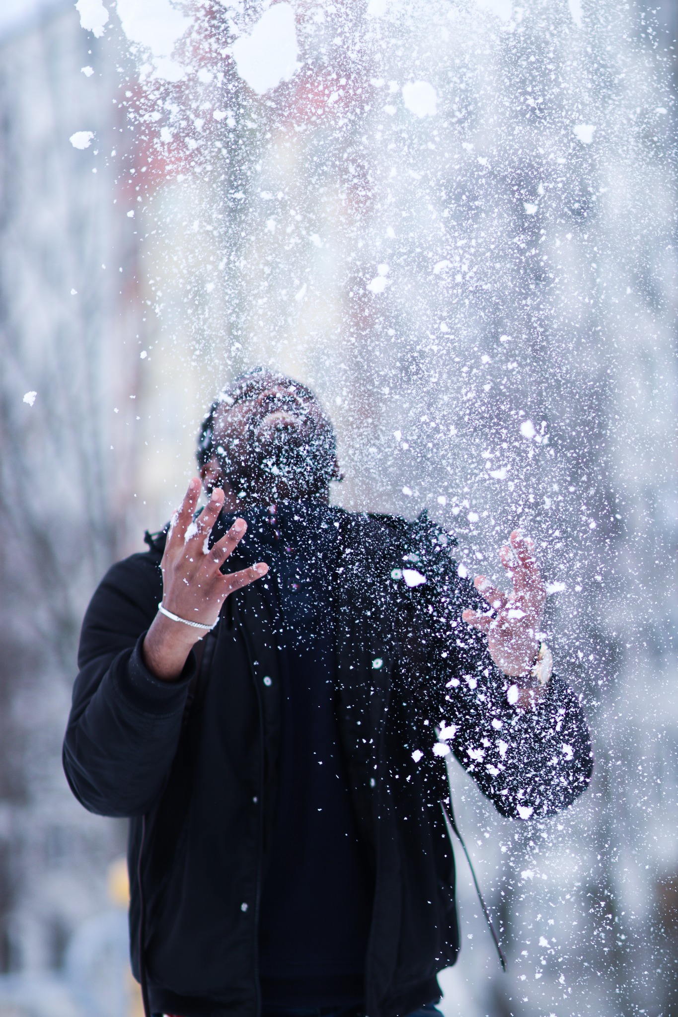 Mężczyzna w czarnej kurtce rzuca śniegiem w górę, zasłaniając twarz chmurą białego puchu, widoczne dłonie z srebrną bransoletką na tle zimowego krajobrazu.