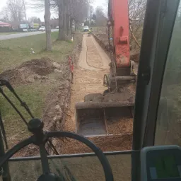 Widok z kabiny koparki na wykopany wąski rów wzdłuż drogi, z widocznymi pracownikami w oddali i drzewami w tle.