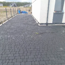 Ułożona, ciemnoszara kostka brukowa na podwórku domu jednorodzinnego, z widocznym wężem ogrodowym i fragmentem ściany budynku.