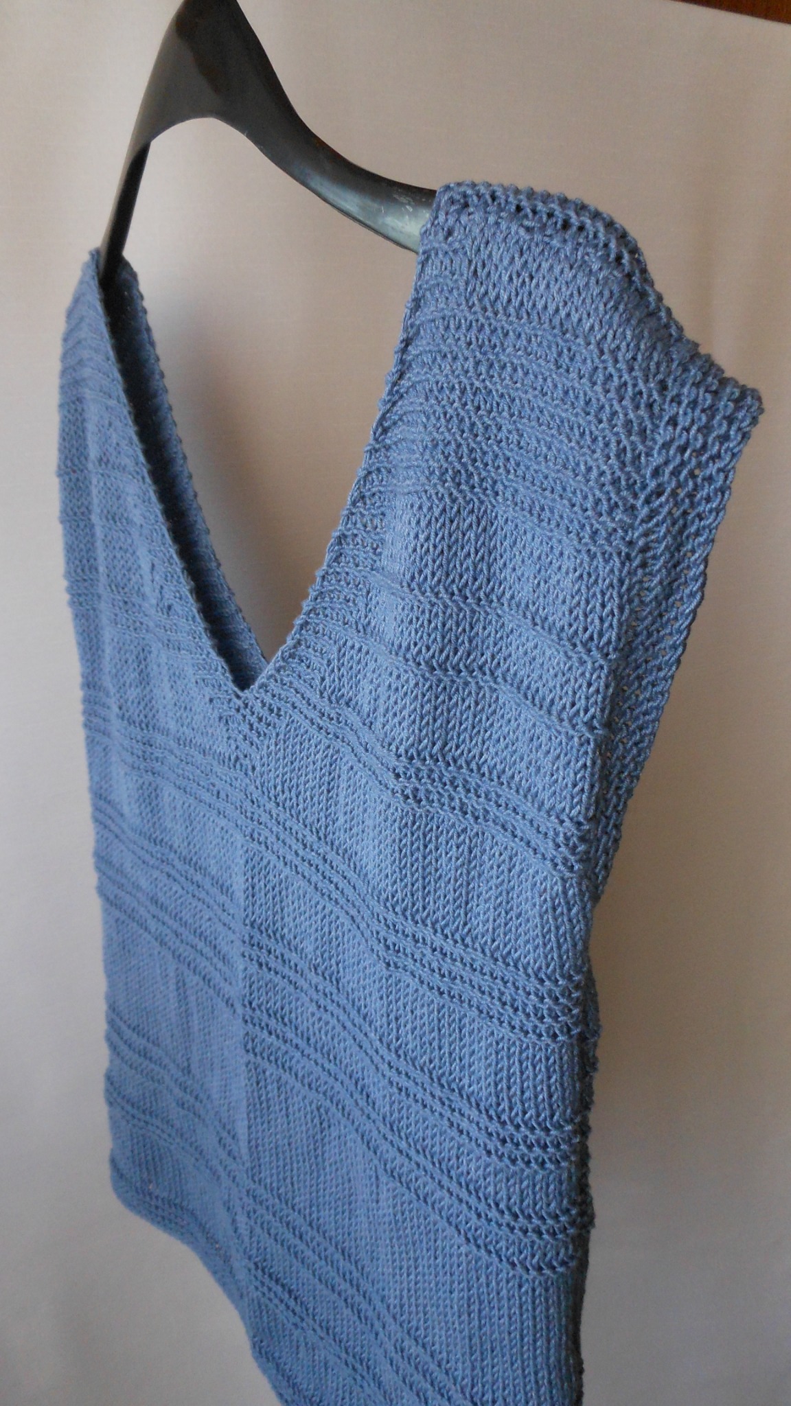 Niebieska, dziergana bluzka z dekoltem w serek, wisząca na czarnym wieszaku, z widoczną fakturą ściegów i poziomymi pasami.