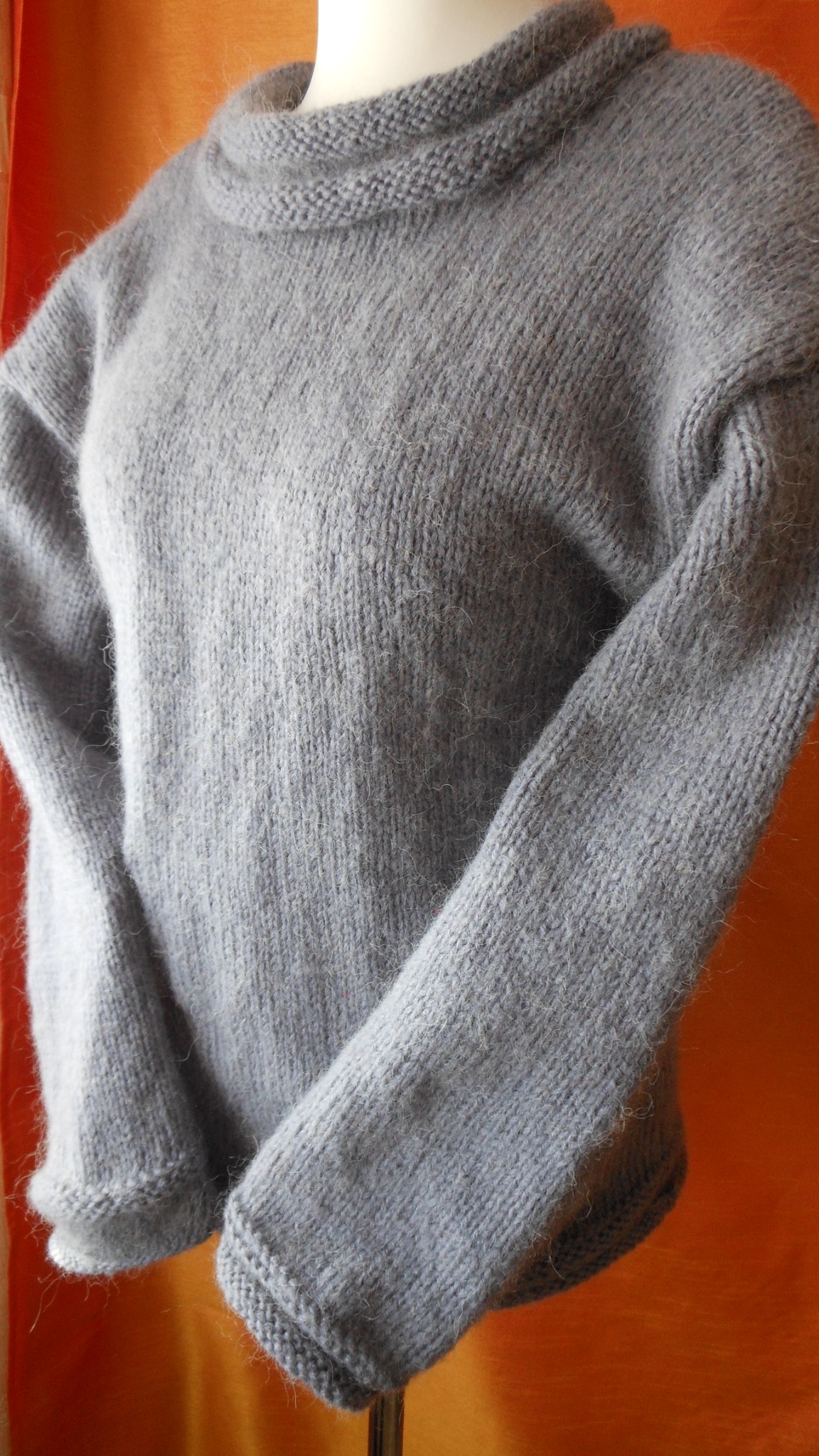 Szary, wełniany sweter z golfem na manekinie, widoczne detale splotu i miękkość włóczki, pomarańczowe tło.