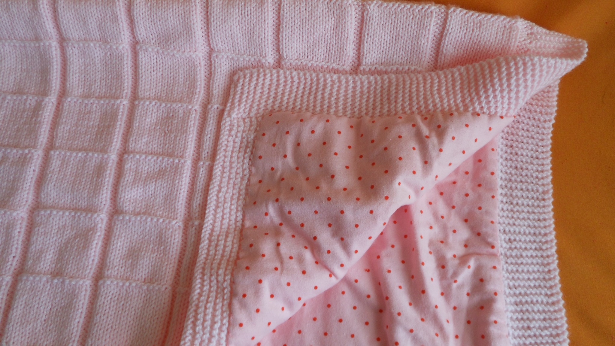 Ręcznie robiony, różowy kocyk z kwadratowym wzorem dzierganym na wierzchu i różową podszewką w czerwone groszki, widoczny fragment złożonego rogu.