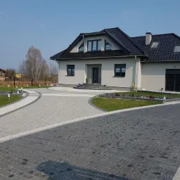 Nowoczesny dom jednorodzinny z jasną elewacją i ciemną dachówką, otoczony brukowanym podjazdem z dekoracyjnym oświetleniem ogrodowym i trawnikami, widok z poziomu gruntu.