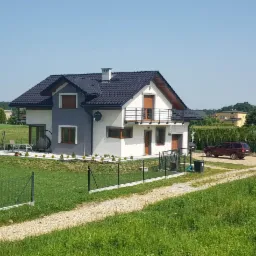 Dwukondygnacyjny dom z szarą i białą elewacją, ciemnym dachem, balkonem z czarną balustradą i małym tarasem, otoczony zielonym trawnikiem i ogrodzeniem z siatki.