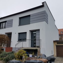 Odnowiona elewacja domu z szarymi panelami dekoracyjnymi i świeżym, jasnym tynkiem. Widoczne schody wejściowe z metalową barierką i fragment ogrodu.