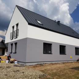 Dwukolorowy dom z szarą elewacją w trakcie malowania. Widoczni pracownicy, puszki z farbą i narzędzia. Tło: pochmurne niebo i sąsiedni budynek.