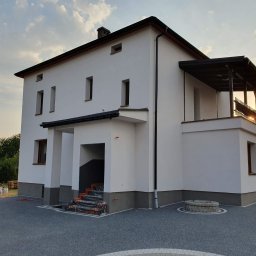 Dwupiętrowy dom z białą elewacją i szarym cokołem, widok z zewnątrz. Na balkonie antena satelitarna, w tle zachodzące słońce. Schody wejściowe zabezpieczone folią.