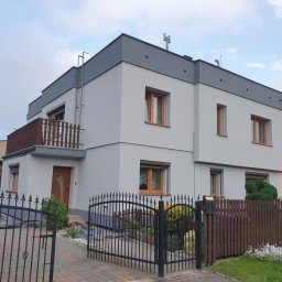 Building Services Sp. z o.o. - Dwukondygnacyjny dom z szarą elewacją i brązowymi oknami, widziany zza czarnego, kutego ogrodzenia. Widoczna drabina na balkonie i fragment sąsiedniego budynku.