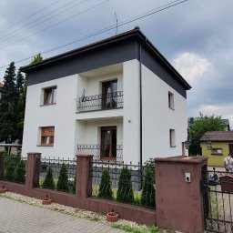 Building Services Sp. z o.o. - Dwukondygnacyjny dom z białą elewacją i ciemnoszarym wykończeniem górnej części, widoczny zza ogrodzenia z brązowymi słupkami i metalowymi przęsłami.