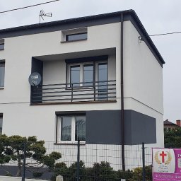 Building Services Sp. z o.o. - Dwukondygnacyjny dom z balkonem, pomalowany na biało i szaro, z anteną satelitarną. Widoczne ogrodzenie i zieleń. Ujęcie z poziomu ulicy w pochmurny dzień.