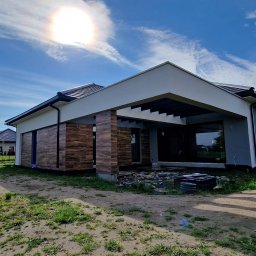 Building Services Sp. z o.o. - Nowoczesny dom jednorodzinny z elementami drewnopodobnej elewacji i szarą elewacją, w trakcie prac wykończeniowych na zewnątrz, w słoneczny dzień.