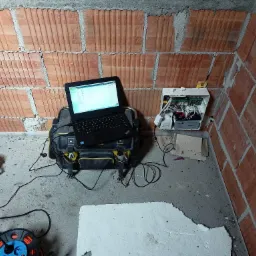 Instalacja alarmowa w surowym wnętrzu: laptop na torbie narzędziowej obok centrali alarmowej na ceglanej ścianie, obok kubek z kawą i przedłużacz.