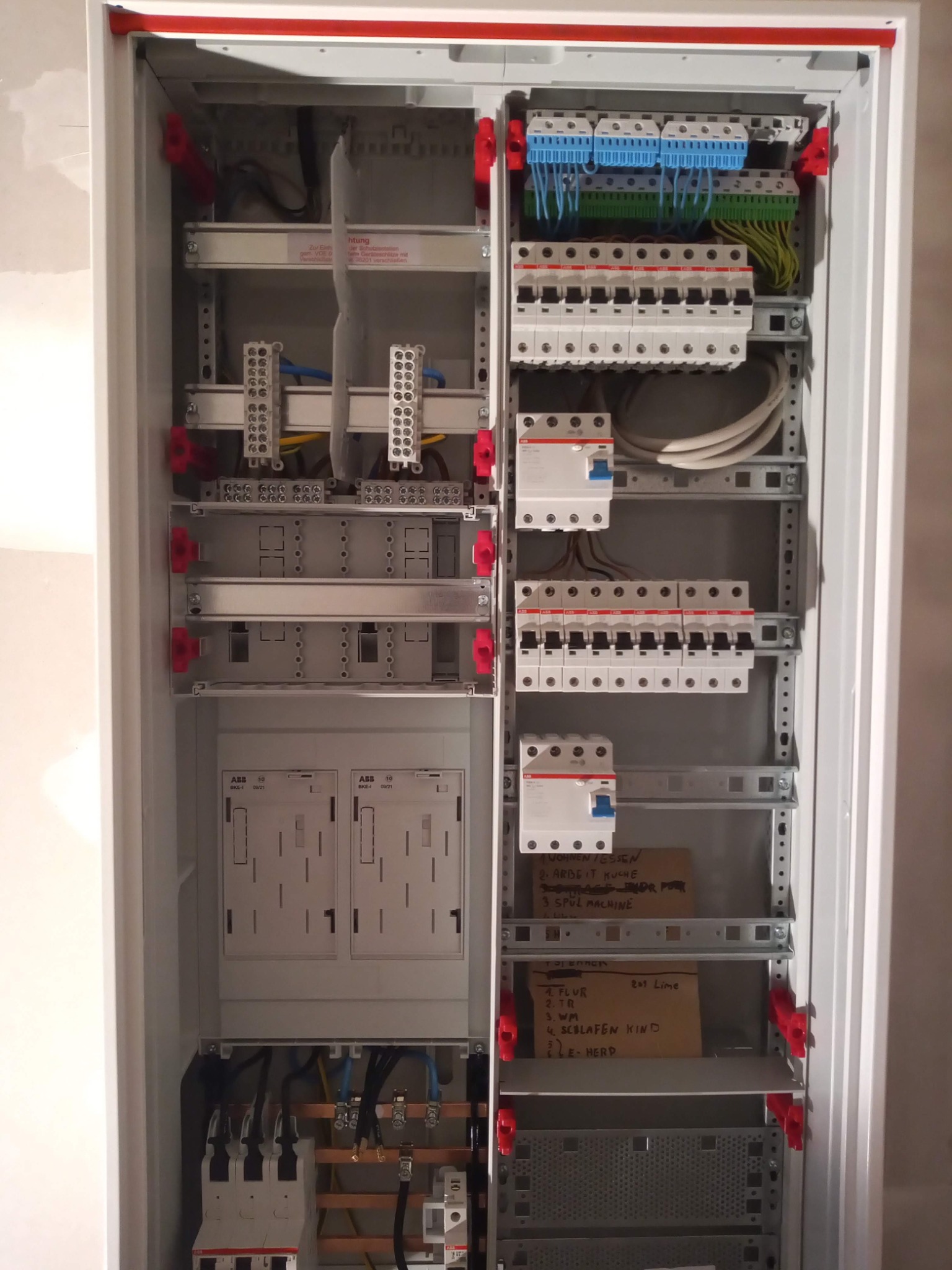 Rozdzielnica elektryczna w białej obudowie, widoczne bezpieczniki, przewody, listwy zaciskowe, moduły ABB, oraz kartki z odręcznymi notatkami opisującymi obwody.