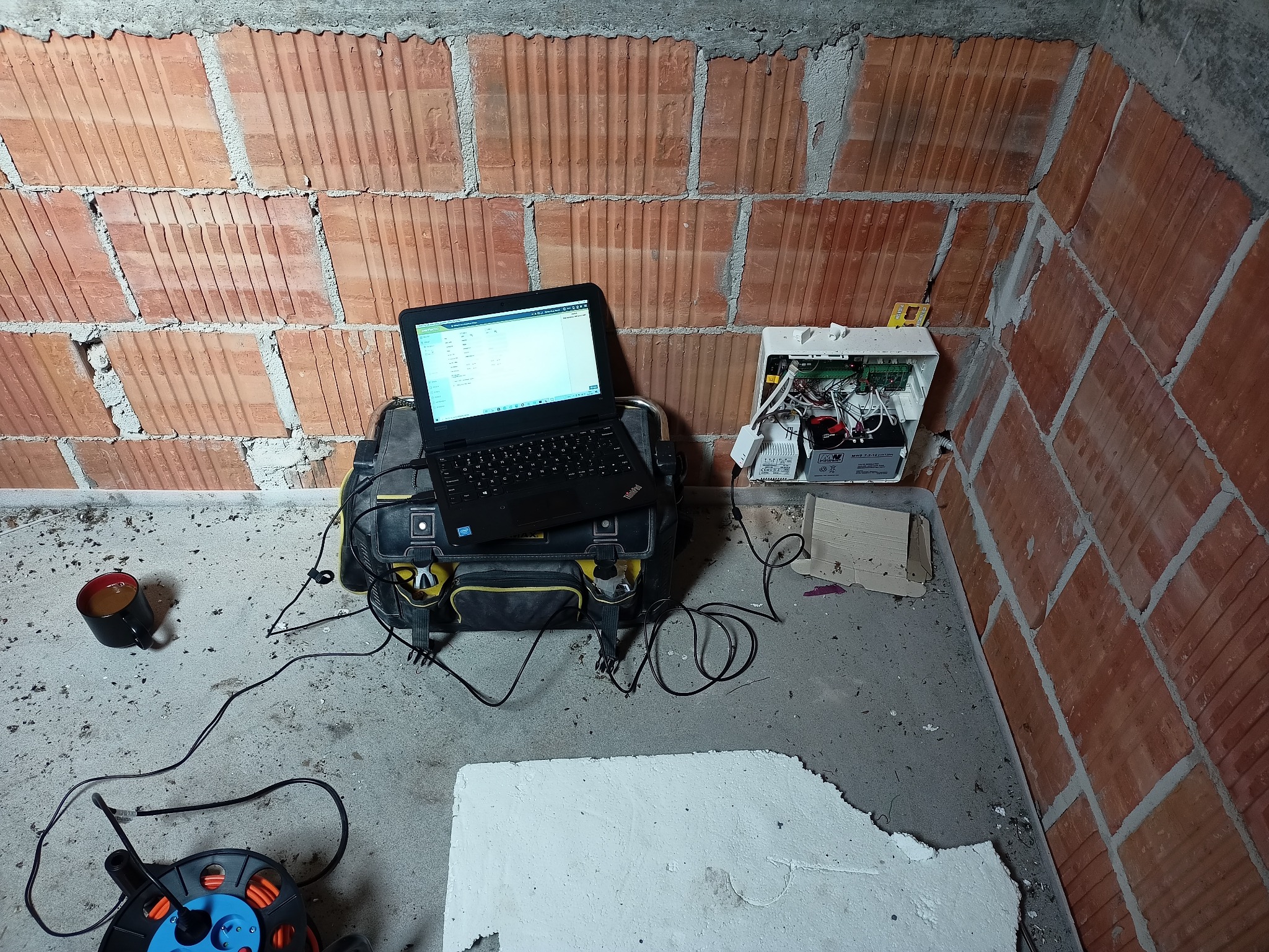 Instalacja alarmowa w surowym wnętrzu: laptop na torbie narzędziowej obok centrali alarmowej na ceglanej ścianie, obok kubek z kawą i przedłużacz.