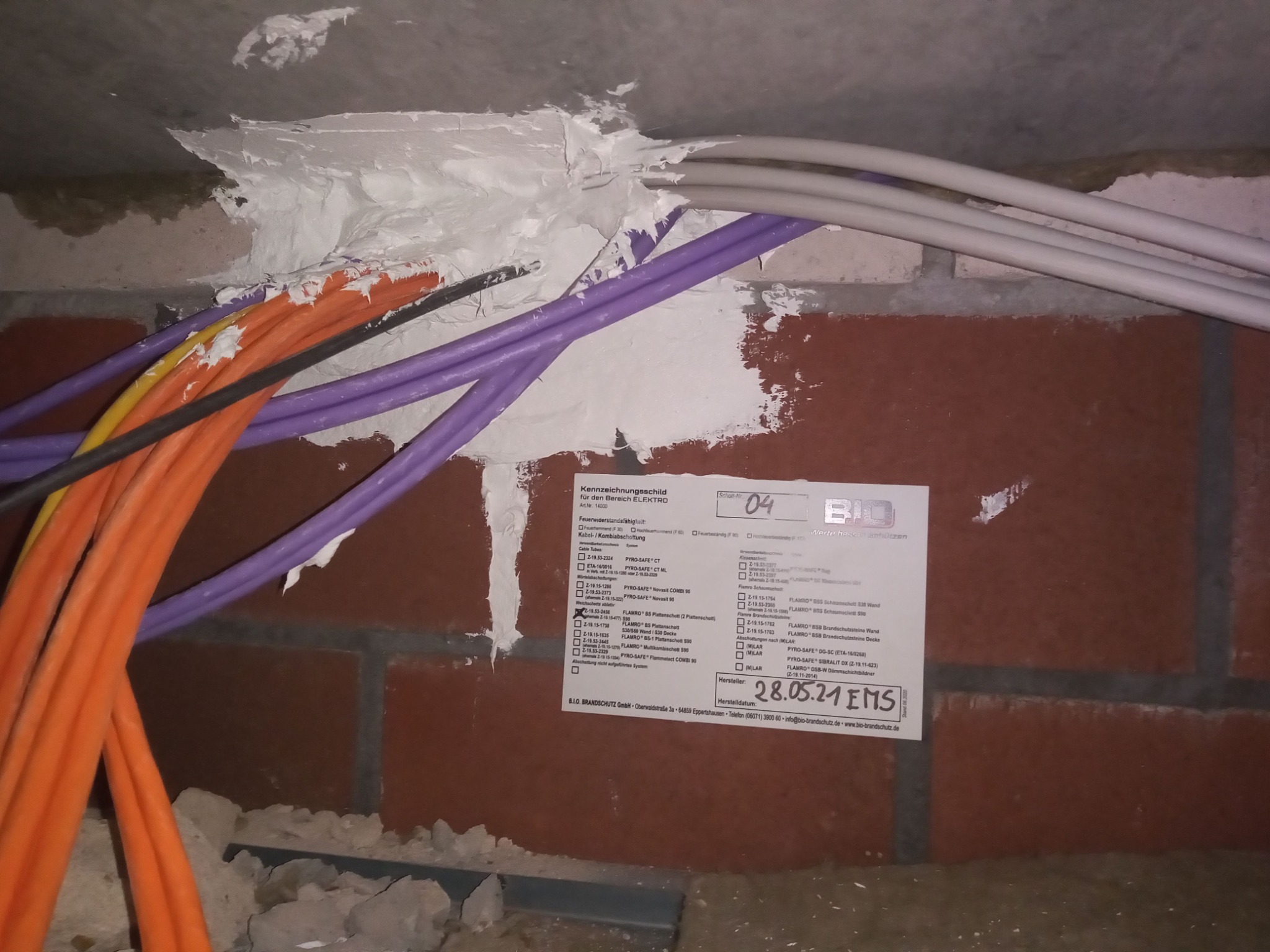 Przejście kabli elektrycznych przez ścianę, zabezpieczone ognioodporną masą uszczelniającą, z naklejoną etykietą identyfikacyjną.