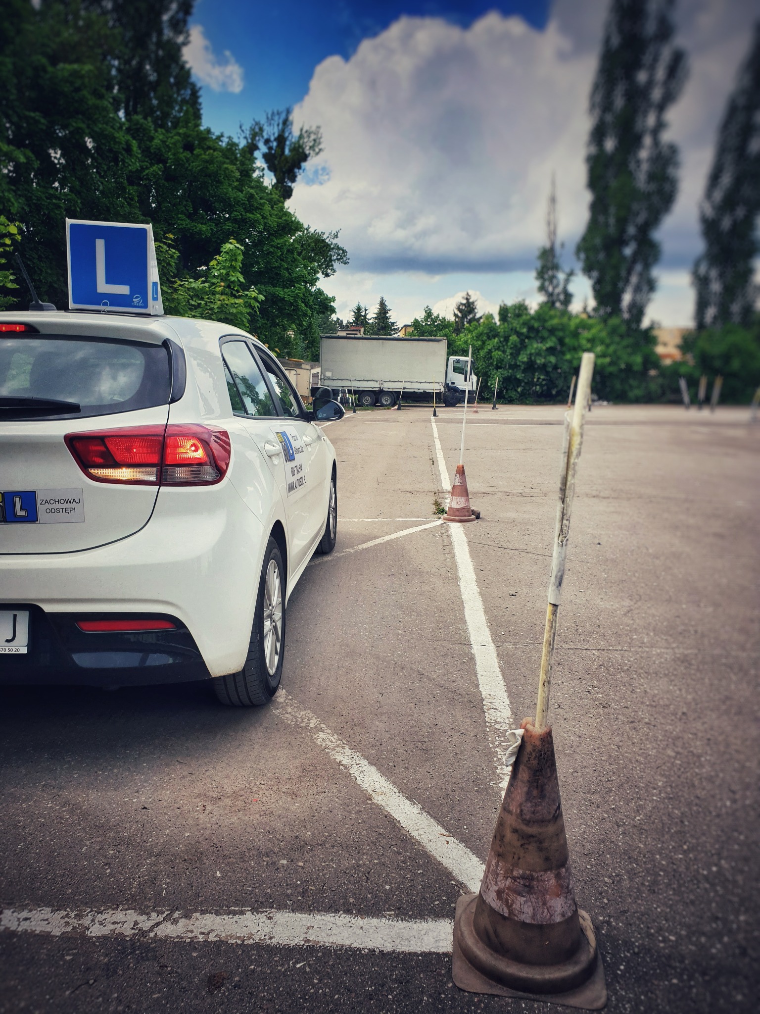 Biały samochód z niebieską literą L na dachu, stojący na placu manewrowym z pachołkami i liniami, w tle drzewa i ciężarówka.
