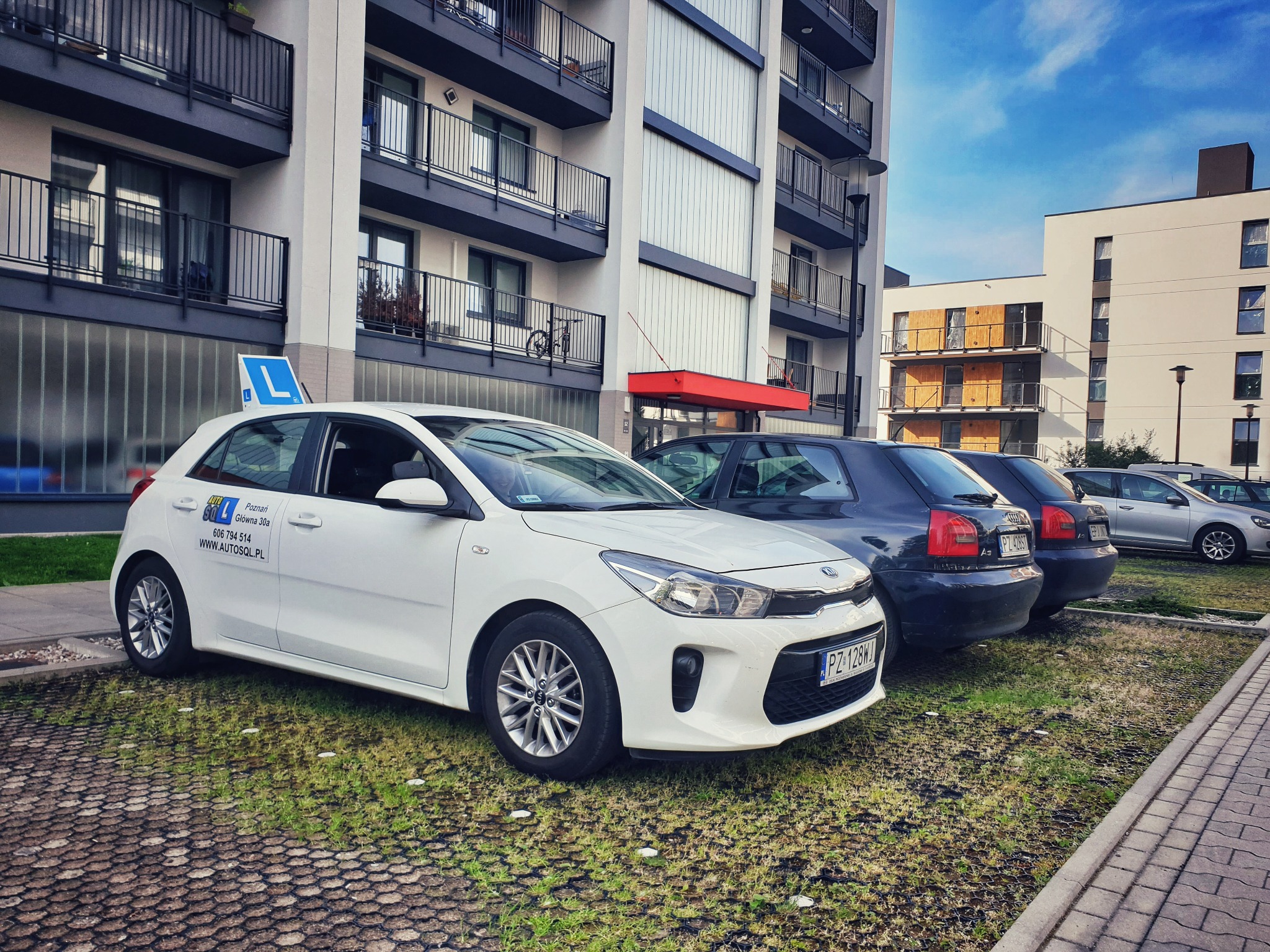 Biały samochód z naklejką i symbolem 'L' na dachu, zaparkowany na trawniku przed nowoczesnym budynkiem mieszkalnym.