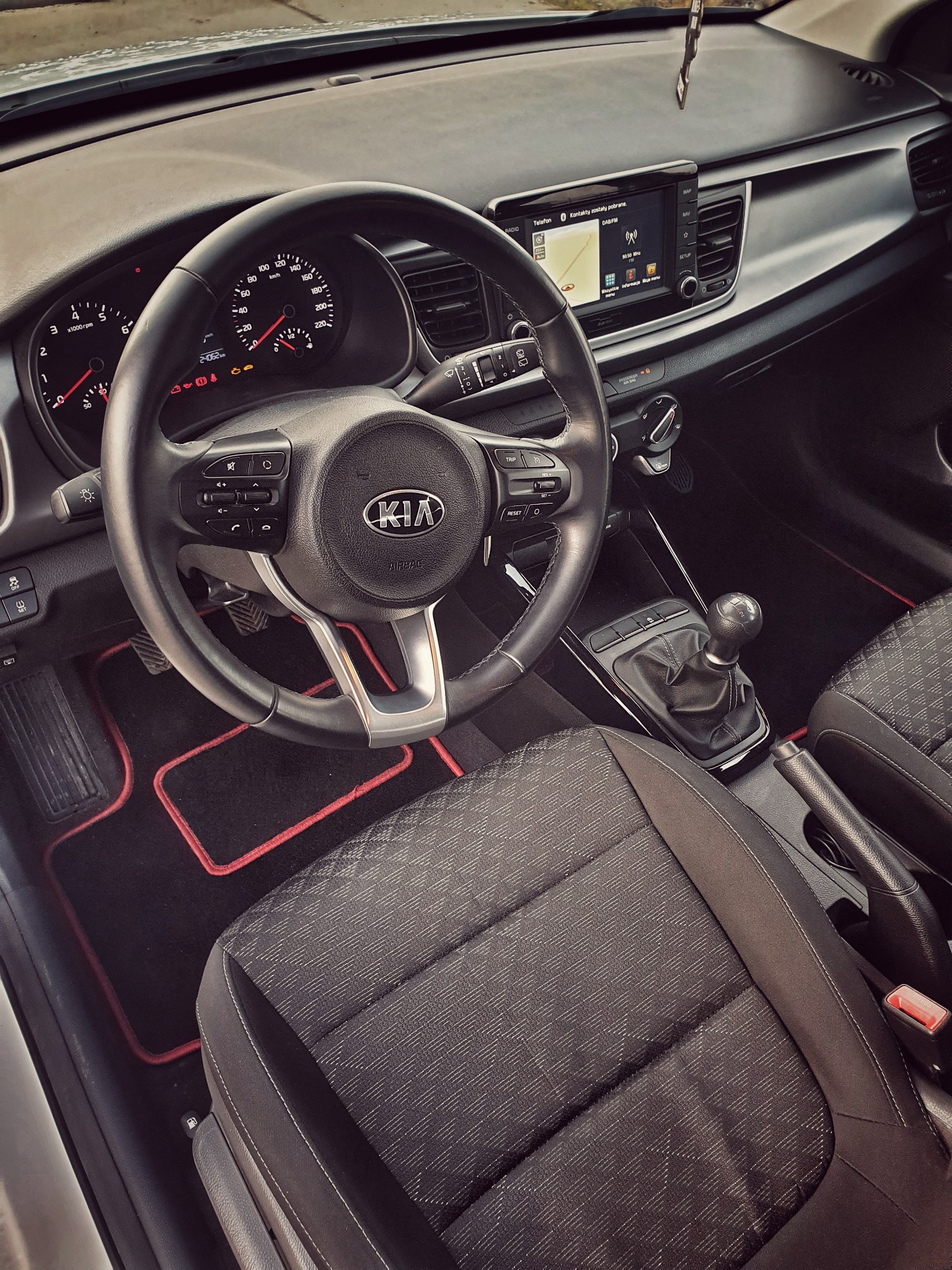 Wnętrze samochodu marki Kia z widoczną deską rozdzielczą, kierownicą z logo, nawigacją na ekranie, dźwignią zmiany biegów oraz czarno-czerwonymi dywanikami.