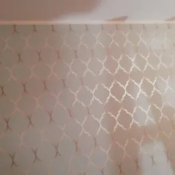 Ściana pokryta tapetą w delikatny, złoty wzór marokański na jasnym, szarozielonym tle, widoczna w narożniku pomieszczenia.