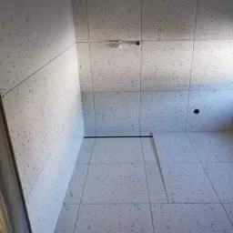 Wykończona łazienka z jasnymi, kwadratowymi płytkami z ciemnymi plamkami na ścianach i podłodze, widoczny odpływ liniowy i niedokończone ułożenie płytek podłogowych.