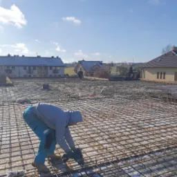 Pracownik w szarym dresie z kapturem, zaginający drut zbrojeniowy na przygotowanej siatce na fundamencie domu, w tle zabudowa jednorodzinna i błękitne niebo.