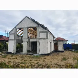 Surowy stan domu w trakcie budowy, ściany z białych bloczków, widoczne drewniane elementy konstrukcji dachu, teren wokół przygotowany pod dalsze prace.