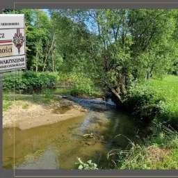 Krajobraz z rzeką i łąką, w tle drzewa, na pierwszym planie logo agencji nieruchomości Fryźlewicz.