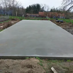 Świeżo wylana, betonowa płyta fundamentowa pod budowę, otoczona ziemią i trawą, widok z góry.