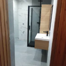 Nowoczesna łazienka z kabiną prysznicową typu walk-in z czarną ramą, umywalką na szafce z drewnianym fornirem i czarną armaturą, wykończona płytkami imitującymi marmur w odcieniach szarości i bieli.