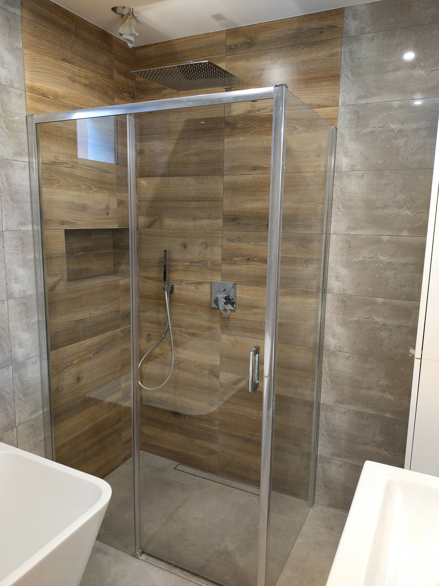 Kabina prysznicowa z deszczownicą i drewnopodobnymi płytkami w nowoczesnej łazience, widoczny fragment umywalki.