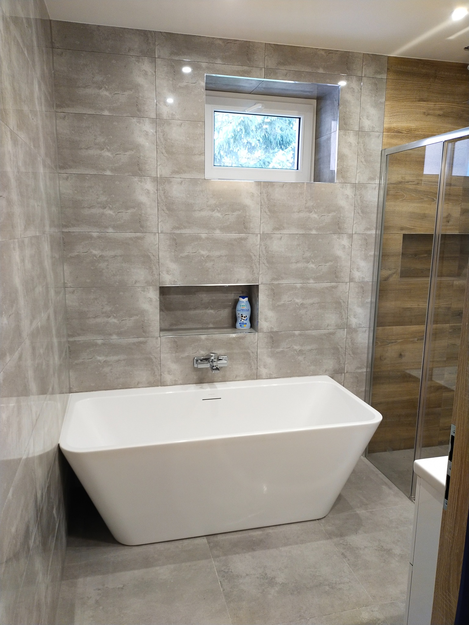 Nowoczesna łazienka z wolnostojącą białą wanną, szarymi płytkami imitującymi kamień na ścianach i podłodze, wnęką z chromowaną baterią i butelką mydła, oraz oknem z widokiem na drzewa.