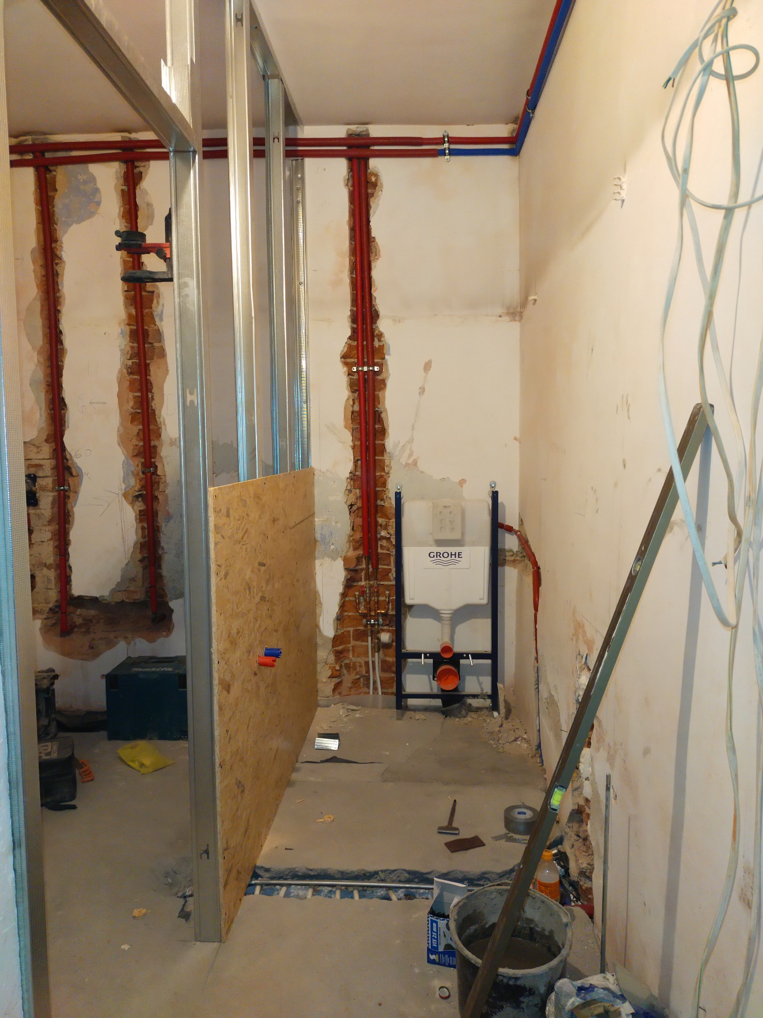 Łazienka w trakcie remontu: widoczne metalowe profile konstrukcyjne, rury instalacji wodnej w ścianach, stelaż podtynkowy WC marki Grohe, płyta OSB, narzędzia i wiadro z zaprawą na podłodze.