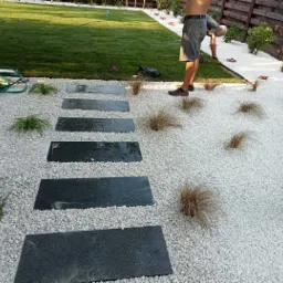 Ścieżka z czarnych płyt kamiennych ułożona na białym żwirze w ogrodzie, z widocznym trawnikiem i roślinami ozdobnymi, oraz pracownikiem ogrodowym w tle.