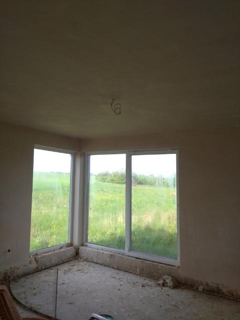 Pokój po nałożeniu świeżych, jasnych tynków; widoczne okna z widokiem na zielone pole, surowe wykończenie podłogi i wystające kable oświetleniowe.