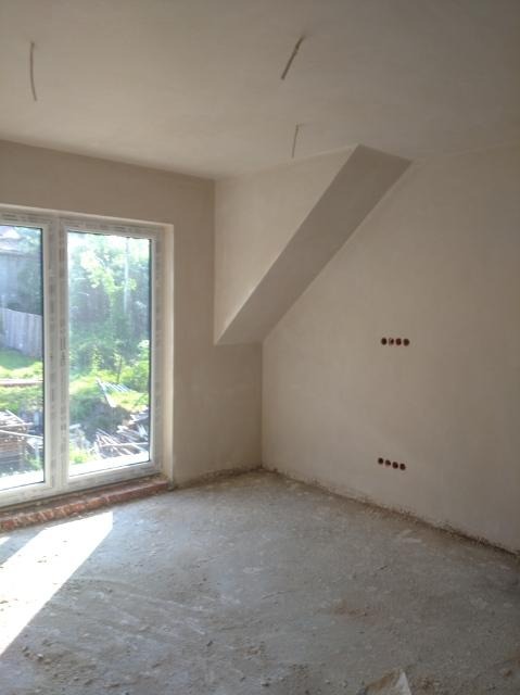 Pokój po tynkowaniu maszynowym, widoczne gładkie ściany, wnęka ze skosem, przygotowane otwory pod gniazdka elektryczne i nieotynkowana podłoga w surowym stanie.