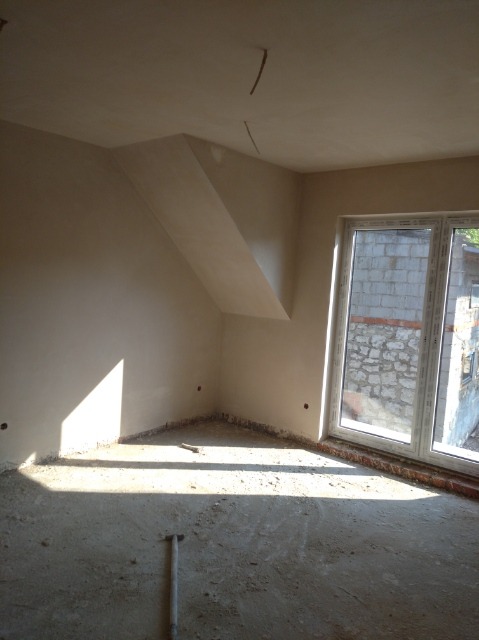 Pokój po nałożeniu gładzi gipsowej, widoczne ślady po instalacji elektrycznej na suficie i narzędzie do wygładzania na podłodze, okno z widokiem na mur z kamienia.