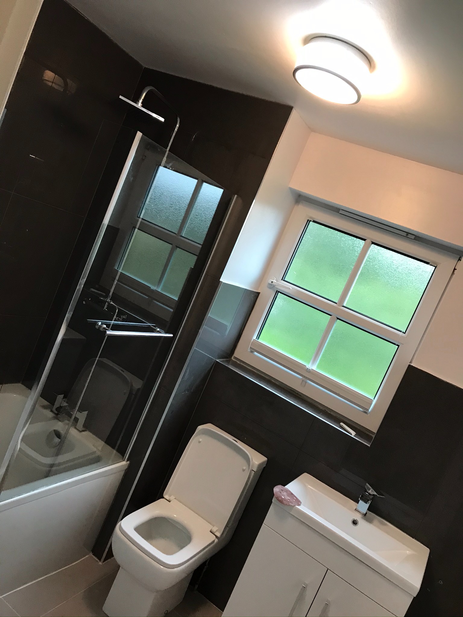 Nowoczesna łazienka z ciemnymi płytkami, kabiną prysznicową, toaletą i białą szafką z umywalką, oświetlona okrągłą lampą sufitową, z oknem z zielonkawą szybą zapewniającym prywatność.
