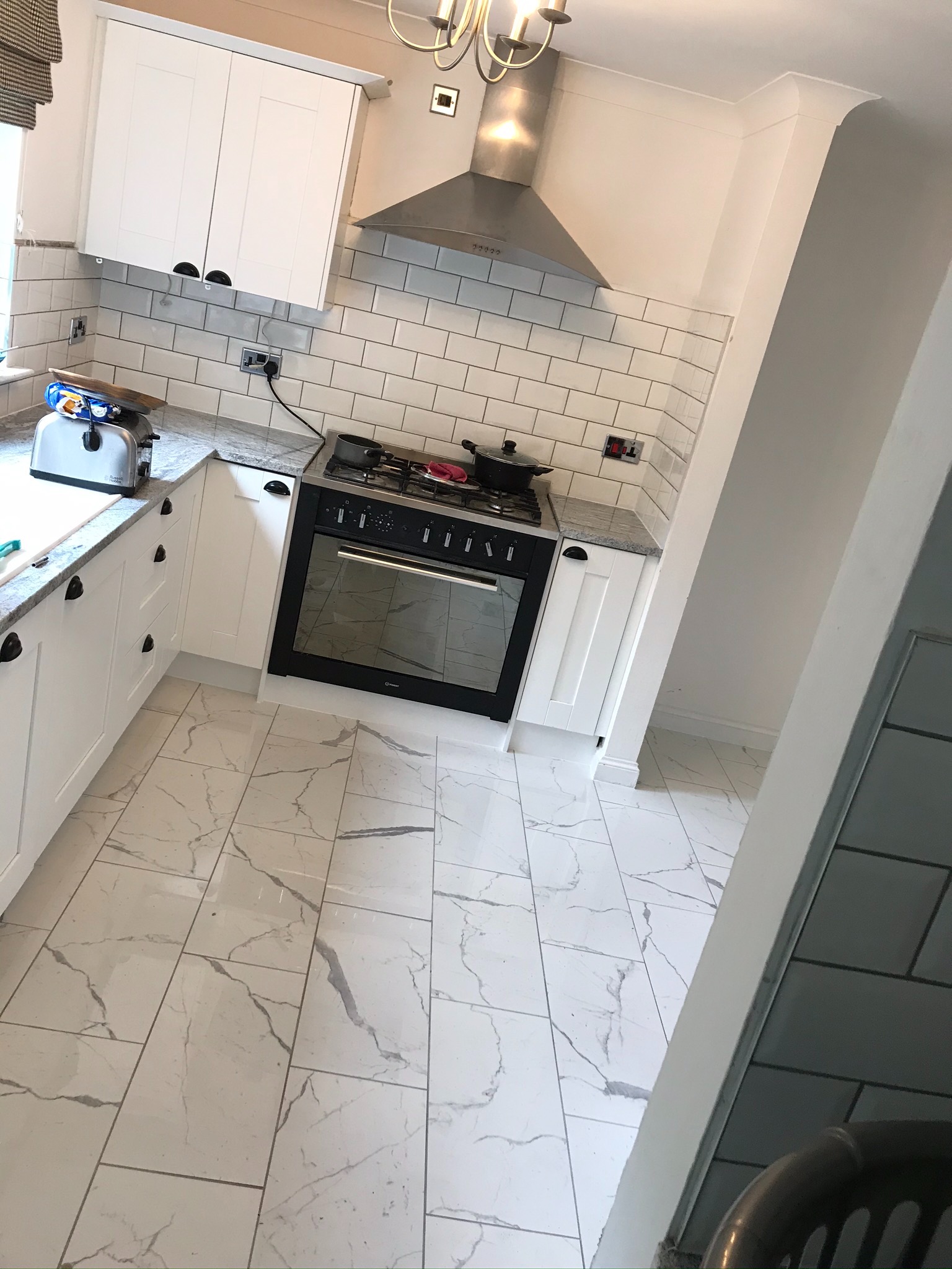 Jasna kuchnia po remoncie: białe szafki z ciemnymi uchwytami, marmurowe płytki podłogowe, białe płytki ścienne ułożone w cegiełkę, srebrny okap i czarna kuchenka.