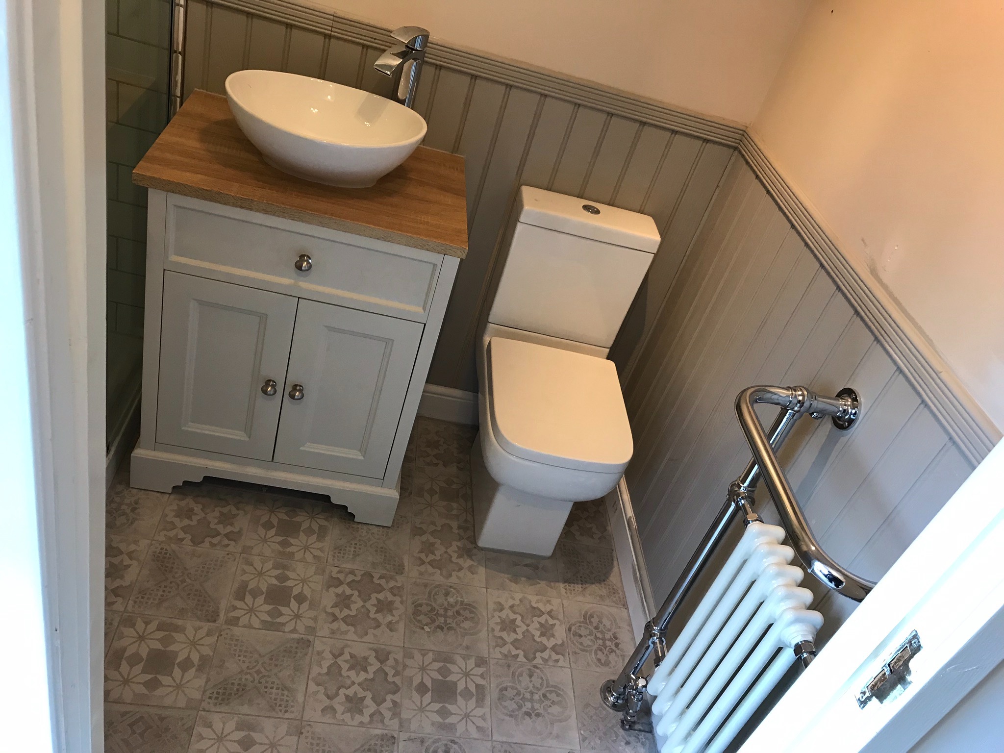 Umywalka nablatowa na szafce z drewnianym blatem, toaleta i grzejnik drabinkowy w odnowionej łazience z szarymi panelami ściennymi i wzorzystą podłogą z płytek.