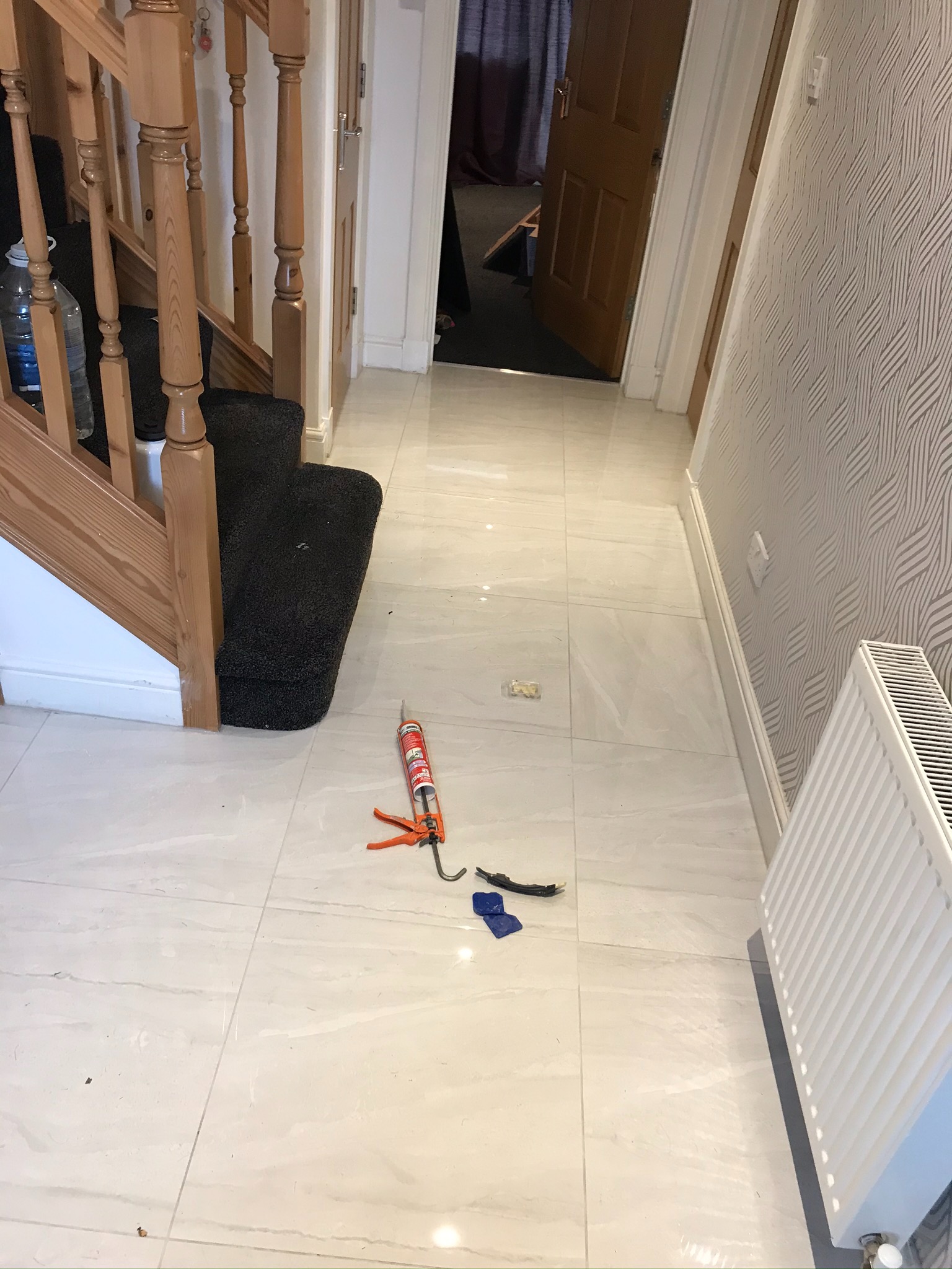 Białe, błyszczące płytki podłogowe w przedpokoju, widoczny aplikator do silikonu, resztki materiałów budowlanych, grzejnik oraz fragment schodów z czarną wykładziną.