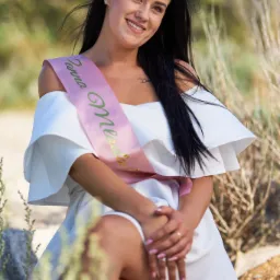 Uśmiechnięta brunetka w białej sukience z odkrytymi ramionami i szarfą z napisem siedzi na kamieniu w plenerze.