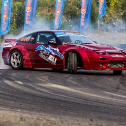 FotoPatrykBierski - Czerwony samochód do driftu z naklejkami sponsorskimi, w trakcie pokazu, z dymem unoszącym się spod kół, na torze z widocznymi śladami opon.